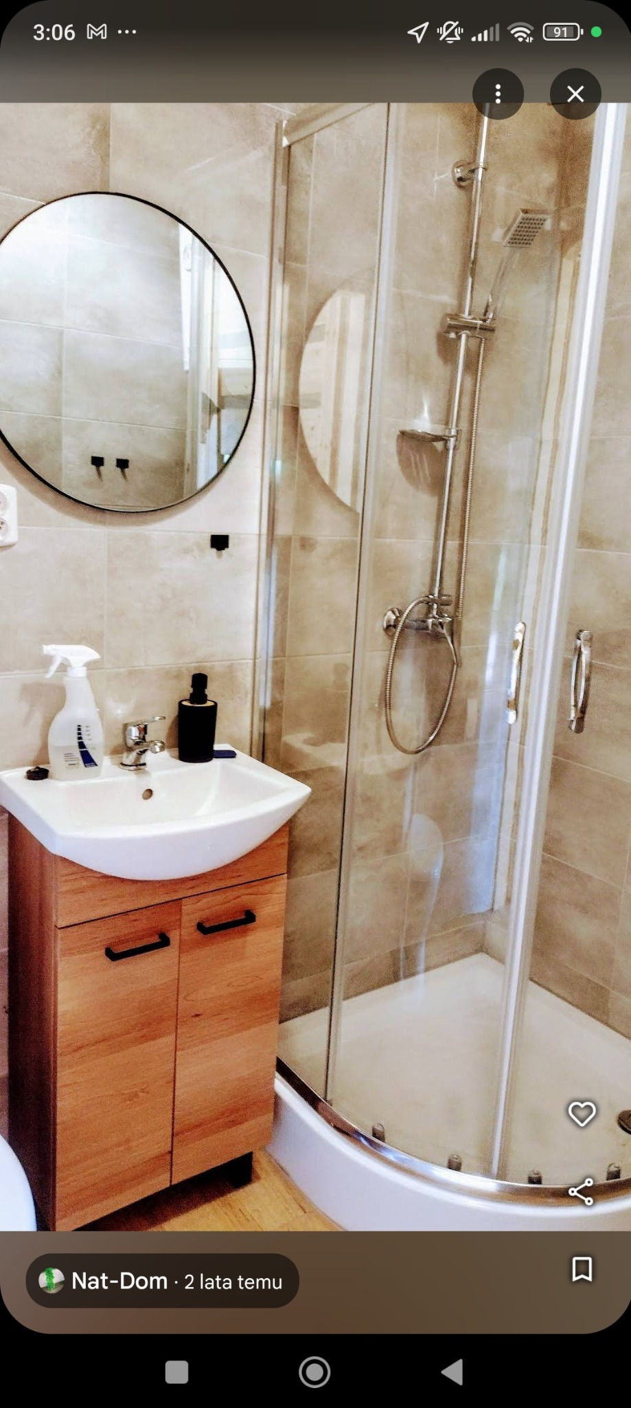 Nowoczesna łazienka z kabiną prysznicową narożną, umywalką nablatową na drewnianej szafce z czarnymi uchwytami, okrągłym lustrem i beżowymi płytkami na ścianach.