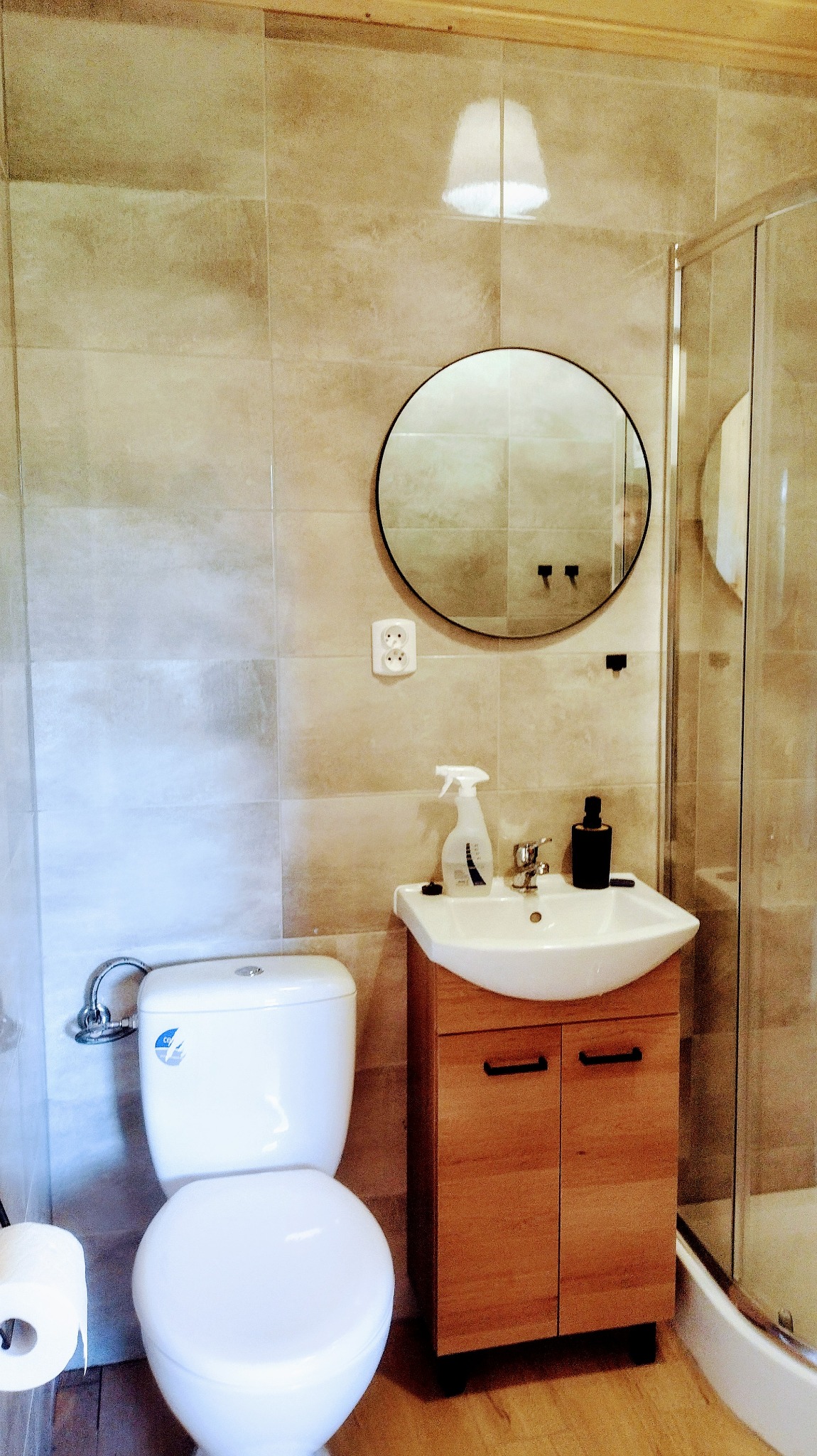 Jasna łazienka z beżowymi płytkami, okrągłym lustrem, białą toaletą i umywalką z szafką z jasnego drewna, widoczny fragment kabiny prysznicowej.