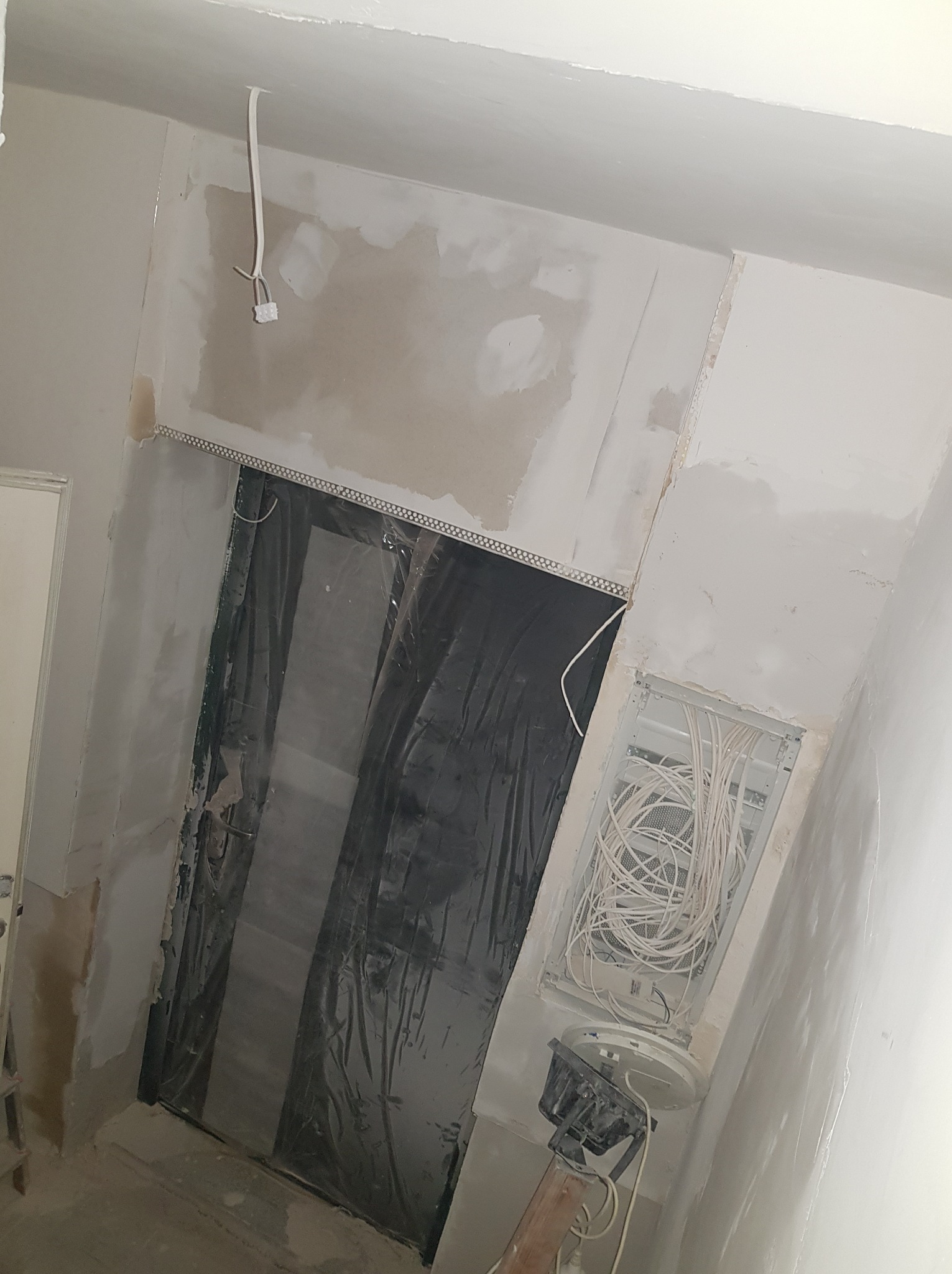 Wnętrze w trakcie remontu: widoczne kable elektryczne w puszce podtynkowej, zabezpieczone folią drzwi, surowe ściany z widocznymi śladami gładzi i wystającym przewodem z sufitu.