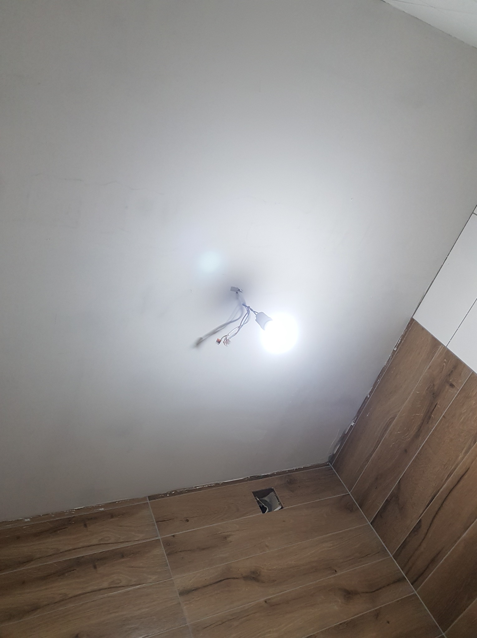 Niedokończona instalacja oświetleniowa na suficie w łazience z brązowymi płytkami imitującymi drewno, widoczne kable i prowizoryczna żarówka.