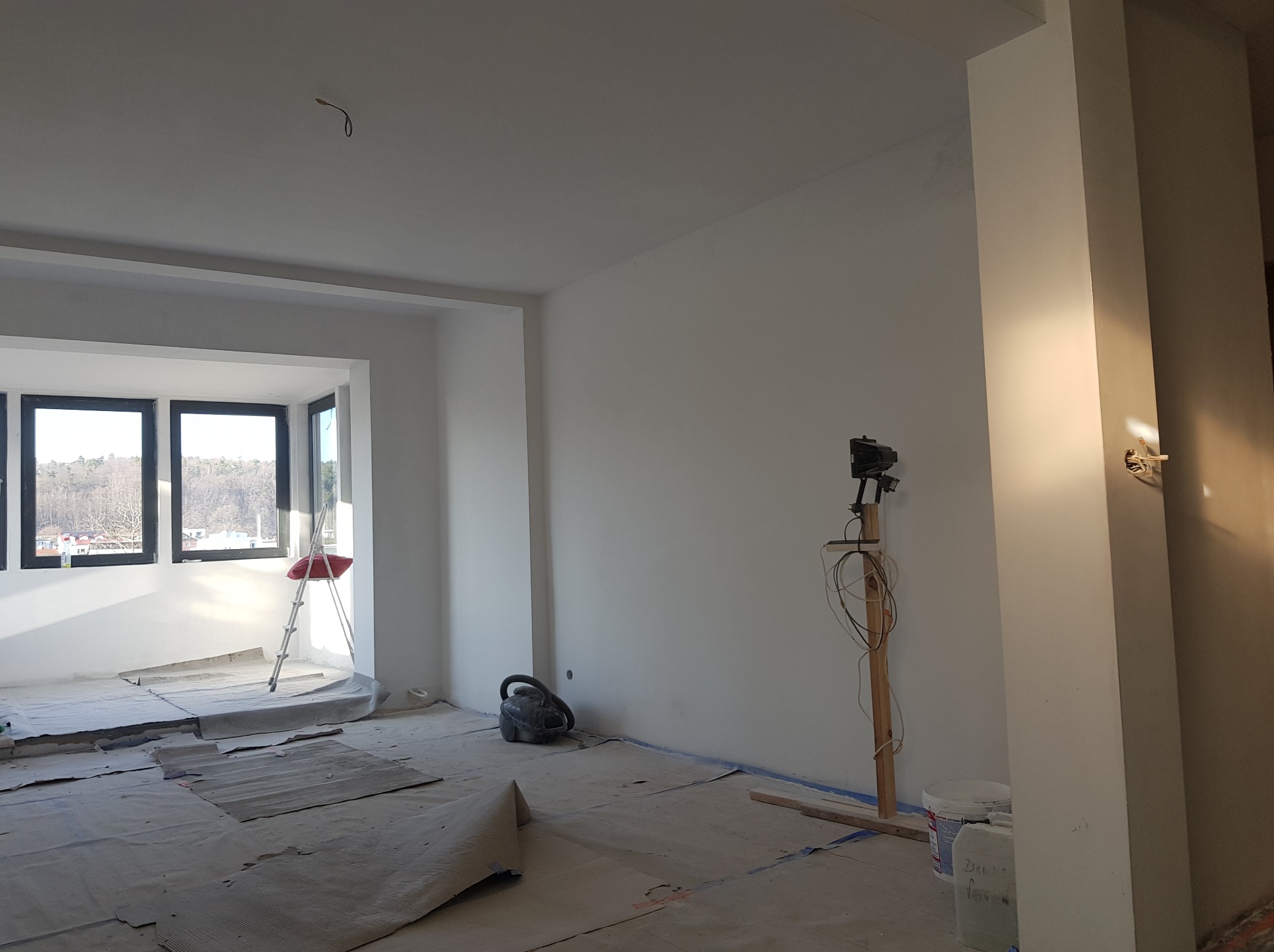 Pokój w trakcie malowania, zabezpieczona podłoga, drabina przy oknie, pistolet do malowania na drewnianym stojaku, wiadro z farbą, widoczne niedociągnięcia w malowaniu ścian.