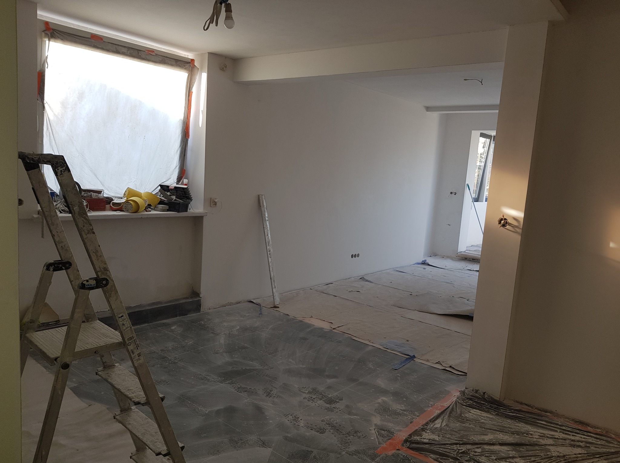 Pokój w trakcie remontu: podłoga zabezpieczona folią, ściany pomalowane na biało, drabina oparta o ścianę, okno zasłonięte folią ochronną, widoczne narzędzia na parapecie.
