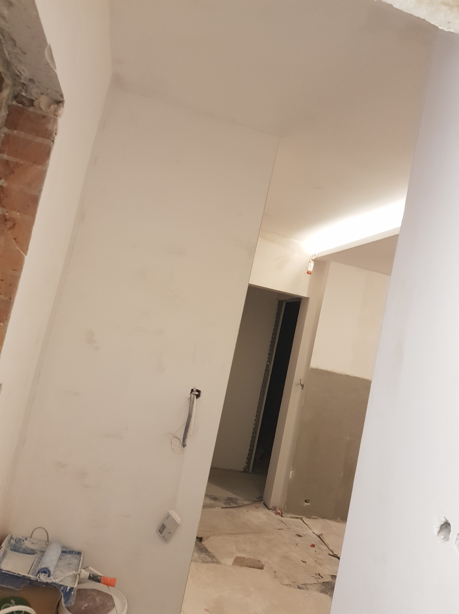 Wnętrze w trakcie remontu: świeżo pomalowane ściany na biało, widoczne narzędzia malarskie w rogu, wystające przewody elektryczne i fragment surowej cegły przy framudze.