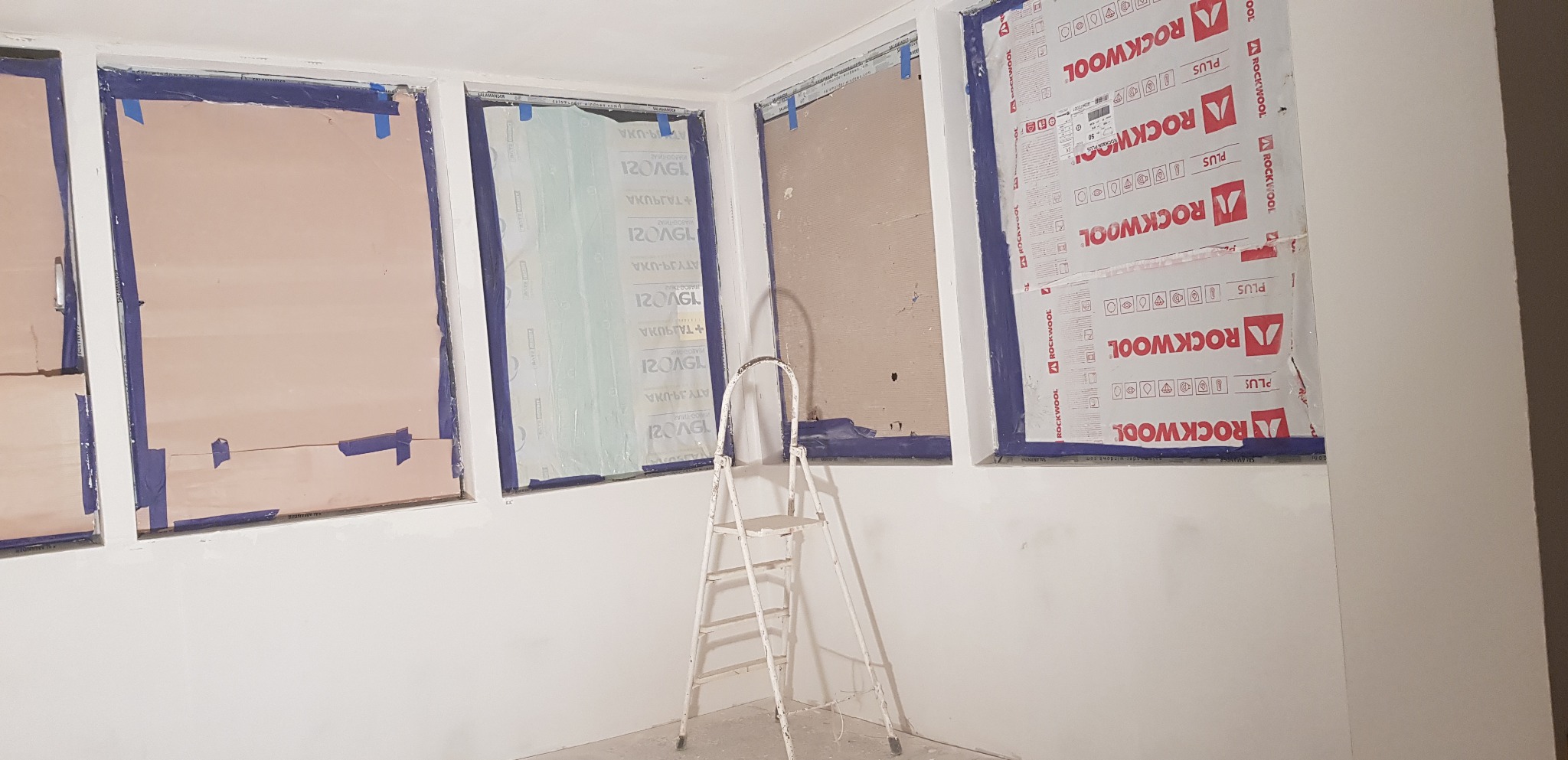 Ocieplanie okien wełną mineralną Rockwool, zabezpieczone taśmą malarską, z widoczną drabinką w surowym, białym pomieszczeniu w trakcie remontu.