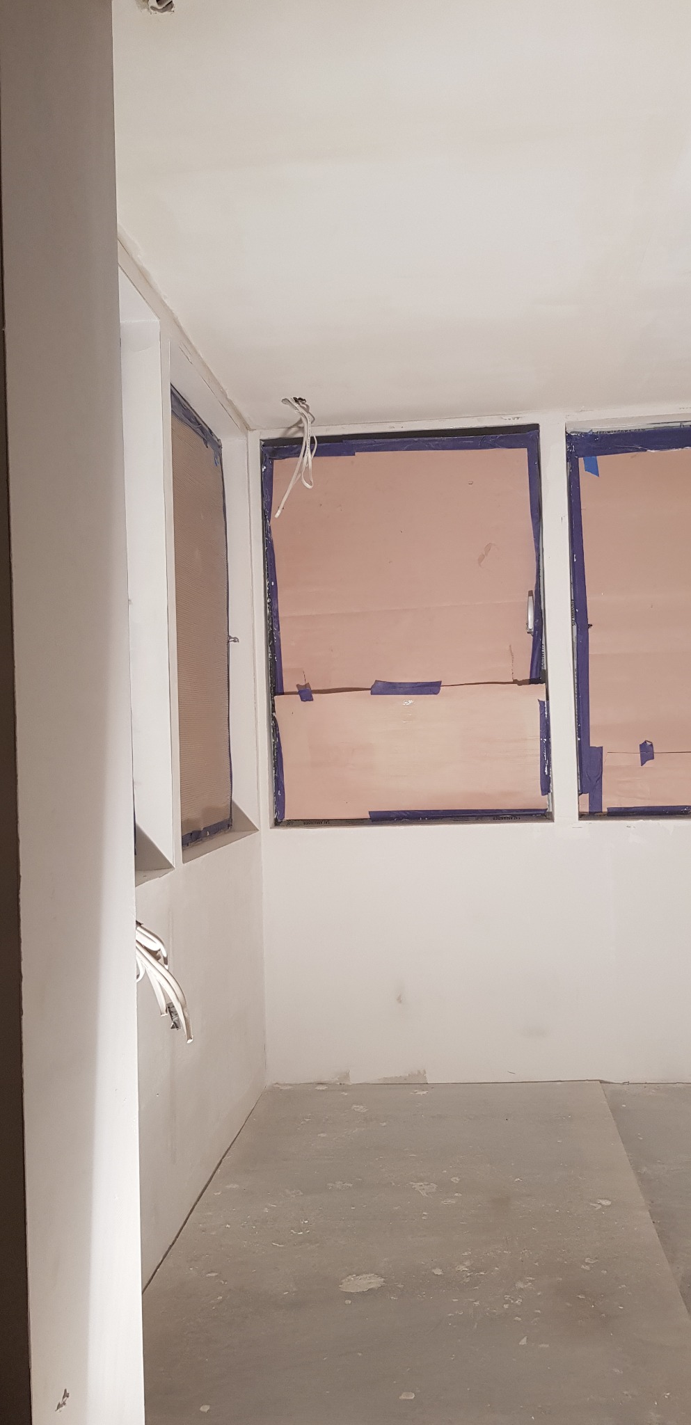 Wnętrze w trakcie remontu: zabezpieczone okna folią malarską, wystające kable elektryczne ze ściany, surowy beton na podłodze, ściany przygotowane do malowania.