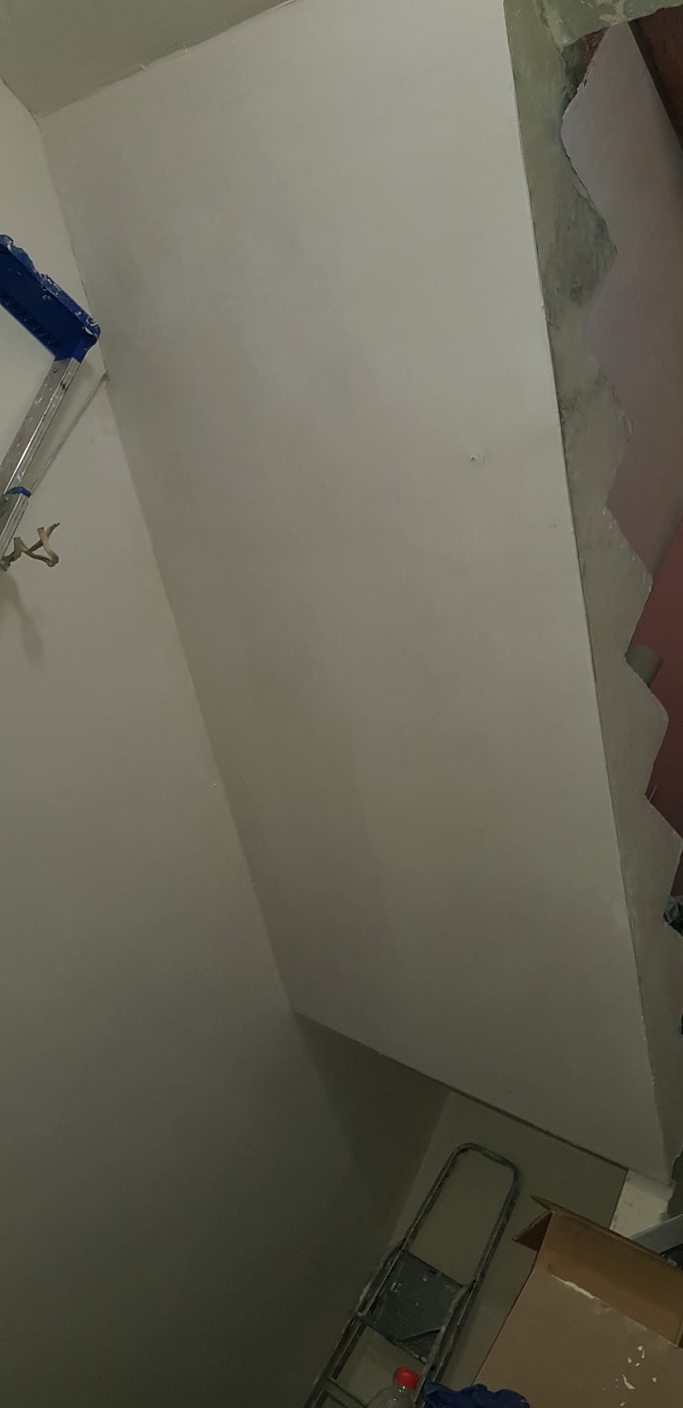 Świeżo pomalowana ściana w narożniku pomieszczenia, widoczny fragment drabiny malarskiej z niebieskim elementem i karton na podłodze, ślady po remoncie na krawędzi ściany.