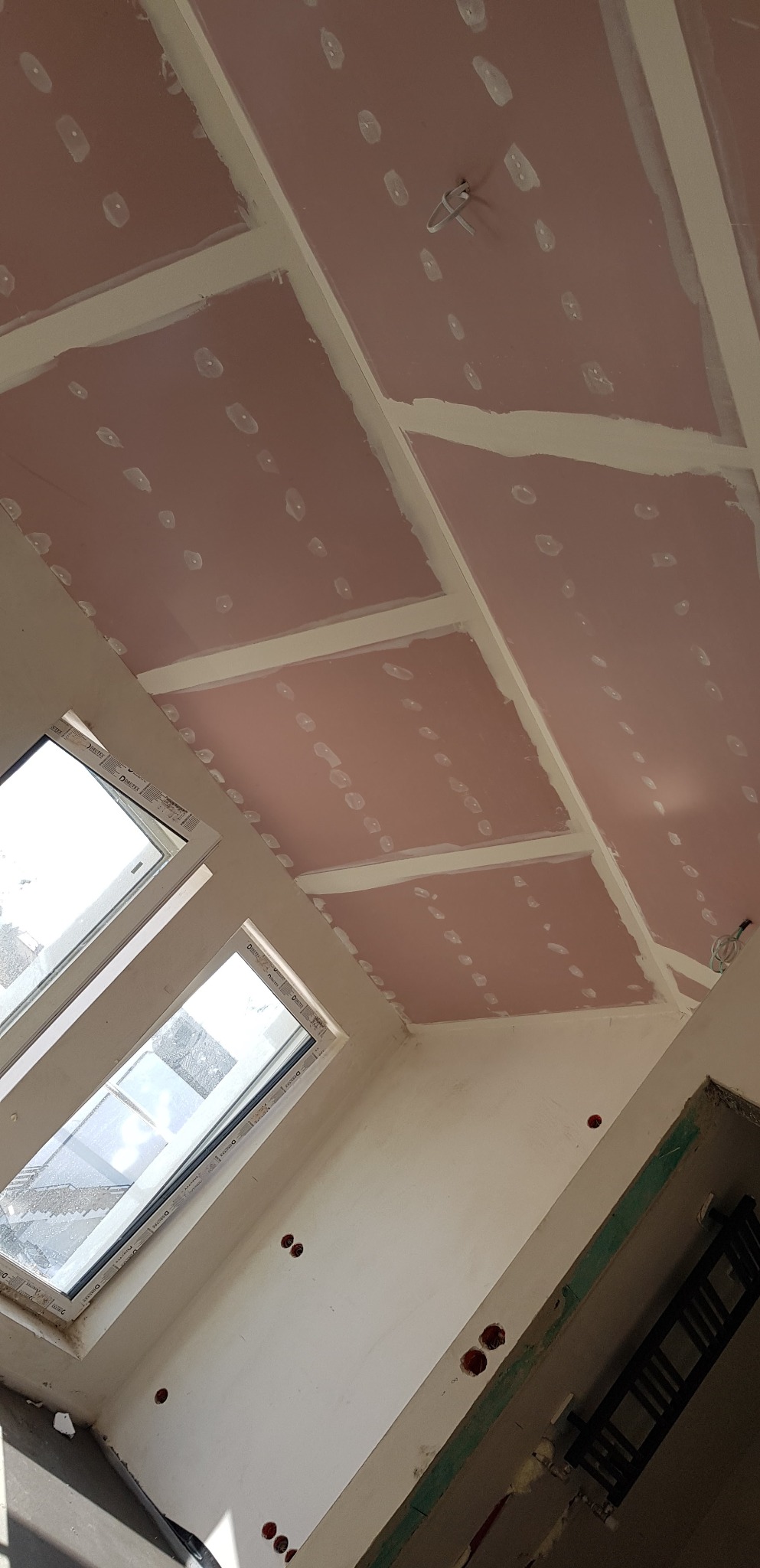 Montaż płyt gipsowo-kartonowych na skośnym suficie poddasza z widocznymi łatami i otworami na punkty oświetleniowe, okna dachowe w tle.