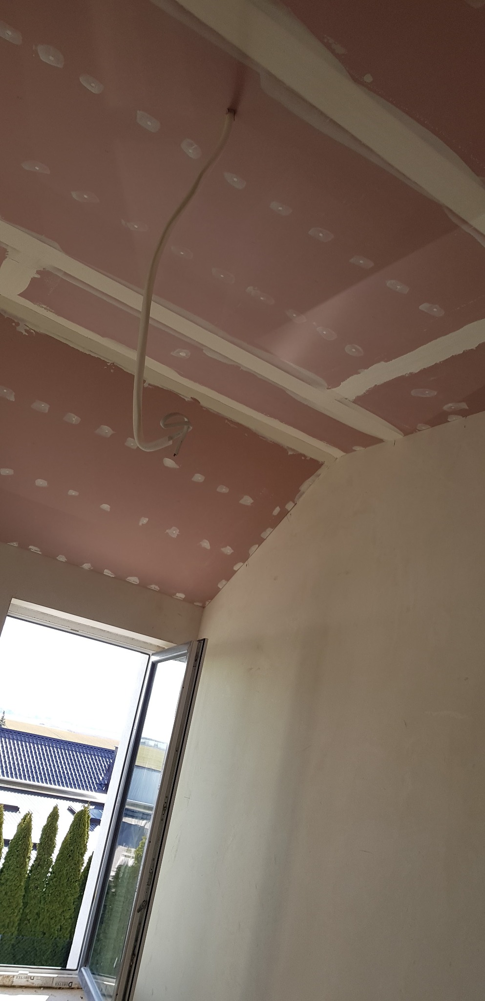 Montaż sufitu podwieszanego z płyt gipsowo-kartonowych z widocznym kablem elektrycznym wystającym z sufitu, na tle otwartego okna z widokiem na zewnątrz.