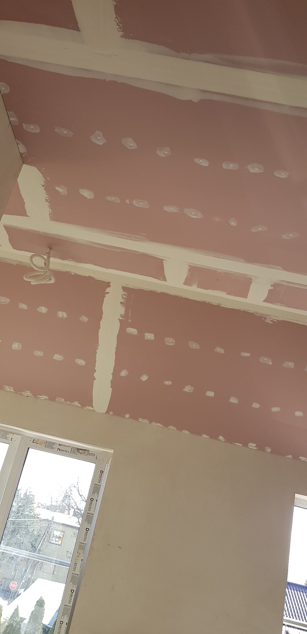 Różowa płyta gipsowo-kartonowa na suficie, z widocznymi spoinami i punktami po wkrętach, przygotowana do dalszych prac wykończeniowych, z fragmentem wystającego okablowania i oknami w tle.