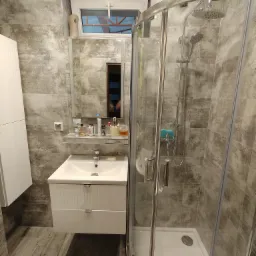 Nowoczesna łazienka z kabiną prysznicową i umywalką w szafce, wykończona płytkami imitującymi beton, z oknem nad lustrem. Wnętrze oświetlone, z widocznymi akcesoriami.