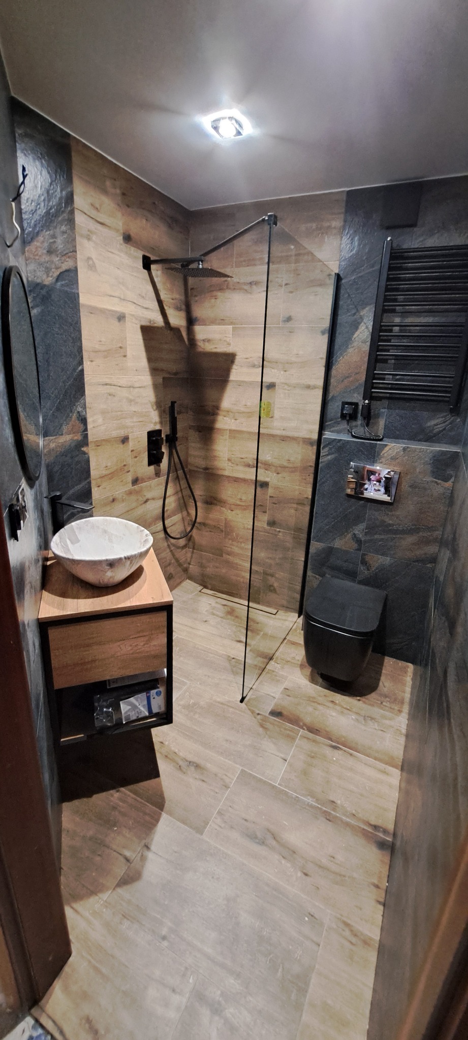 Nowoczesna łazienka z kabiną prysznicową typu walk-in, umywalką nablatową na drewnianej szafce, wiszącą toaletą w kolorze czarnym oraz grzejnikiem drabinkowym, wykończona płytkami imitującymi...