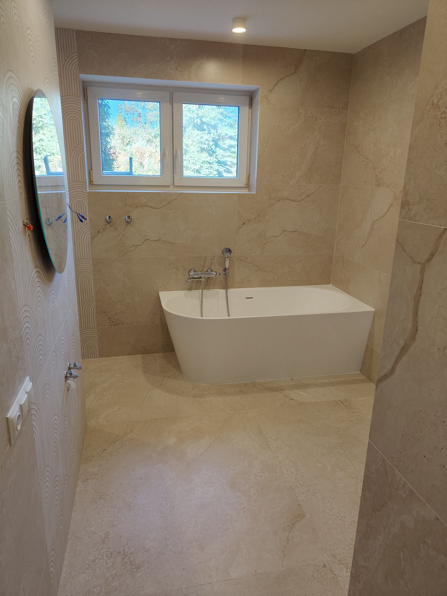 Wykończona łazienka w Wilga z beżowymi płytkami imitującymi marmur, owalnym lustrem i białą wanną prostokątną z baterią. Widoczne okno z widokiem na drzewa.