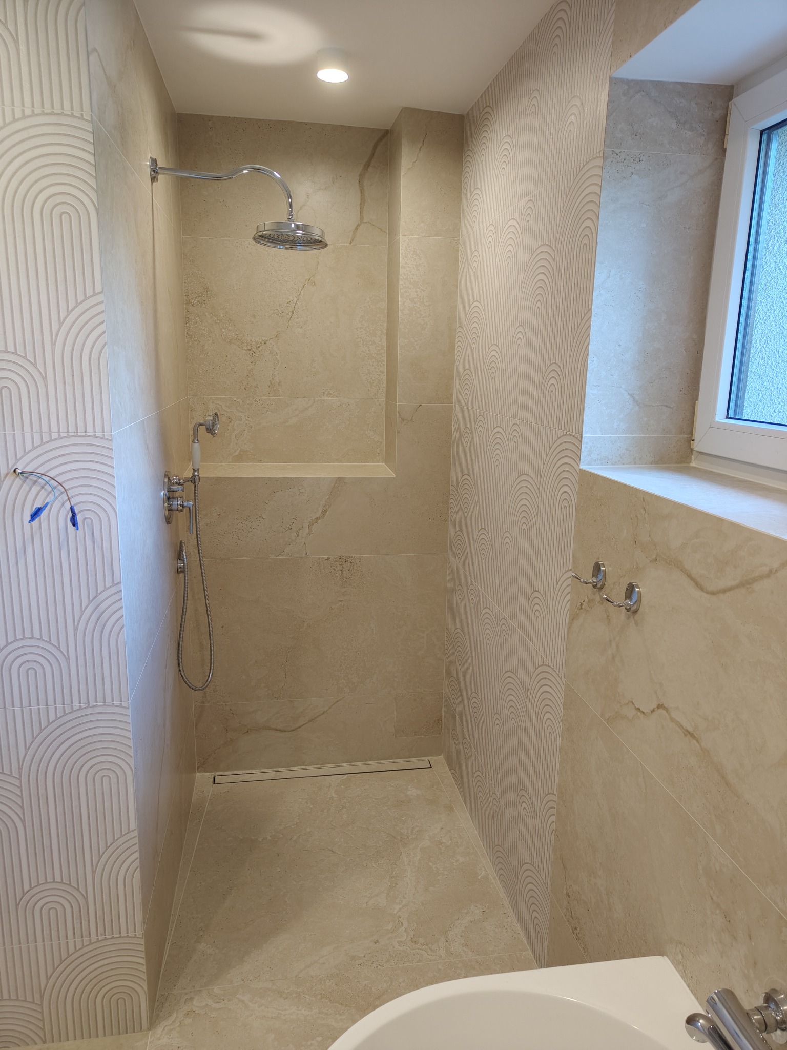 Beżowa kabina prysznicowa z deszczownicą, wykończona płytkami imitującymi marmur oraz płytkami dekoracyjnymi z motywem łuków, widoczne podłączenie do baterii i odpływ liniowy w podłodze.
