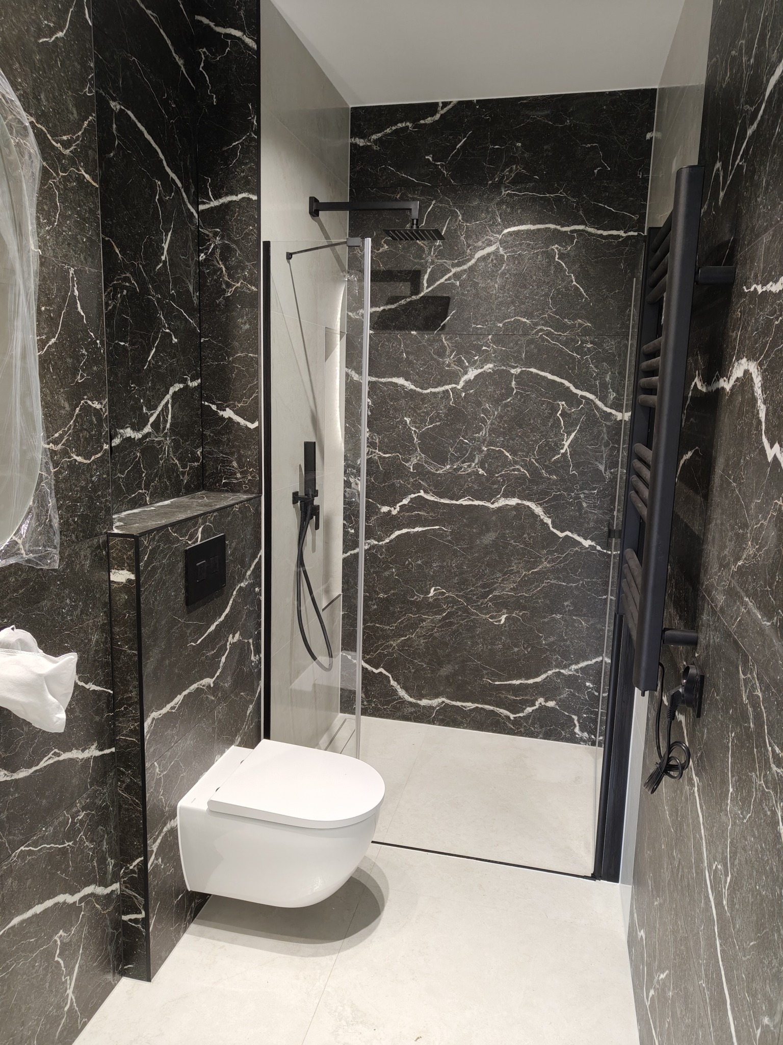 Elegancka łazienka z czarnymi płytkami imitującymi marmur, białą armaturą i kabiną prysznicową ze szklaną ścianką. Grzejnik drabinkowy w kolorze czarnym.