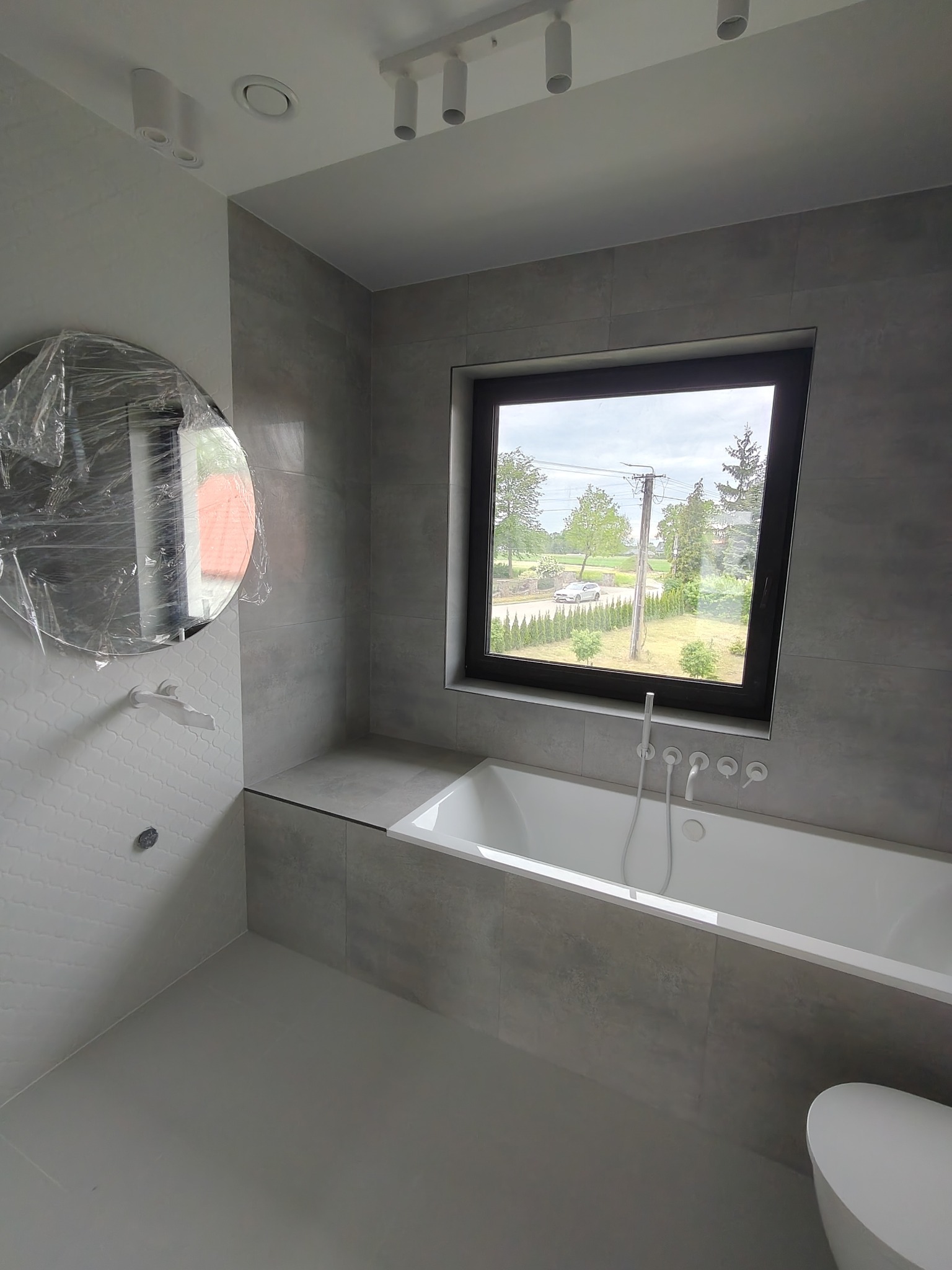 Szara łazienka po remoncie z wanną pod oknem i okrągłym lustrem w folii ochronnej. Widok na zewnątrz przez okno.