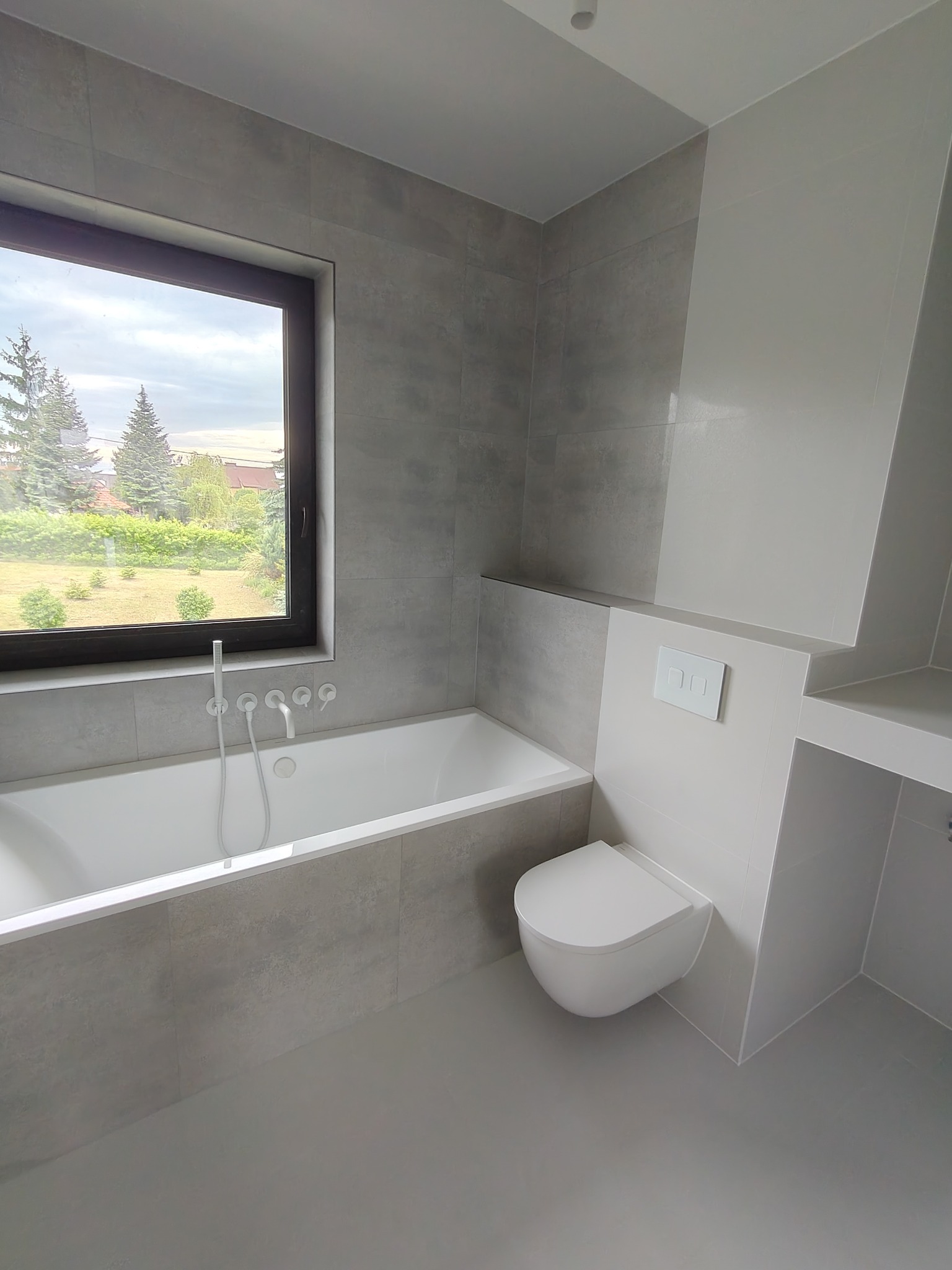 Minimalistyczna łazienka z wanną przy oknie i podwieszaną toaletą. Szare płytki imitujące beton, biała armatura. Widok z okna na zieleń.