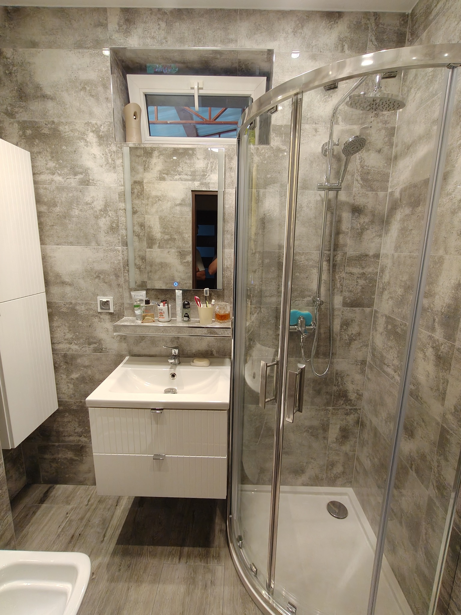 Nowoczesna łazienka z kabiną prysznicową i umywalką w szafce, wykończona płytkami imitującymi beton, z oknem nad lustrem. Wnętrze oświetlone, z widocznymi akcesoriami.