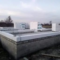 Fundament domu w trakcie budowy: bloczki betonowe na fundamencie, białe pustaki H+H czekające na ułożenie, widok na częściowo zabudowany teren i niebo o zmierzchu.