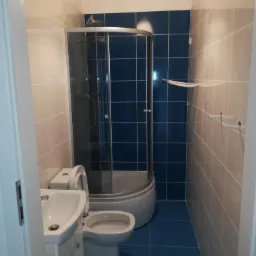 Niewielka łazienka z kabiną prysznicową, umywalką i toaletą, wykończona niebiesko-beżowymi płytkami ceramicznymi z widocznym oświetleniem sufitowym