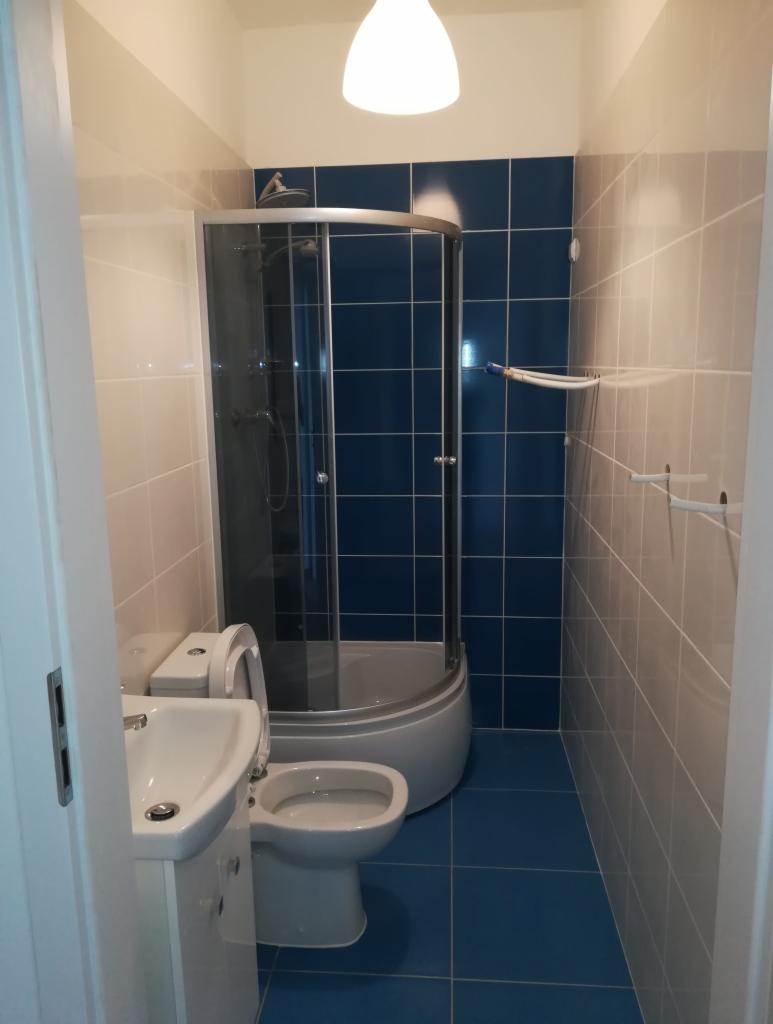 Niewielka łazienka z kabiną prysznicową, umywalką i toaletą, wykończona niebiesko-beżowymi płytkami ceramicznymi z widocznym oświetleniem sufitowym