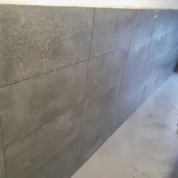 Ściana wyłożona szarymi, matowymi płytkami imitującymi kamień, z widocznymi śladami po kleju na podłodze i elementami instalacji elektrycznej na górze.