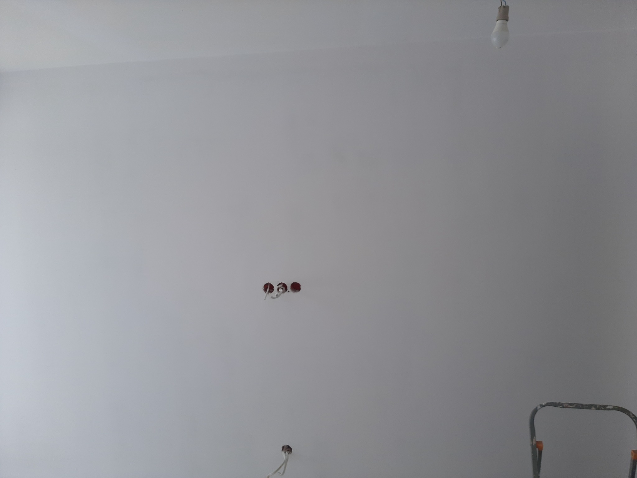 Świeżo pomalowana na biało ściana z wystającymi przewodami elektrycznymi, widoczna drabina malarska i żarówka na suficie.