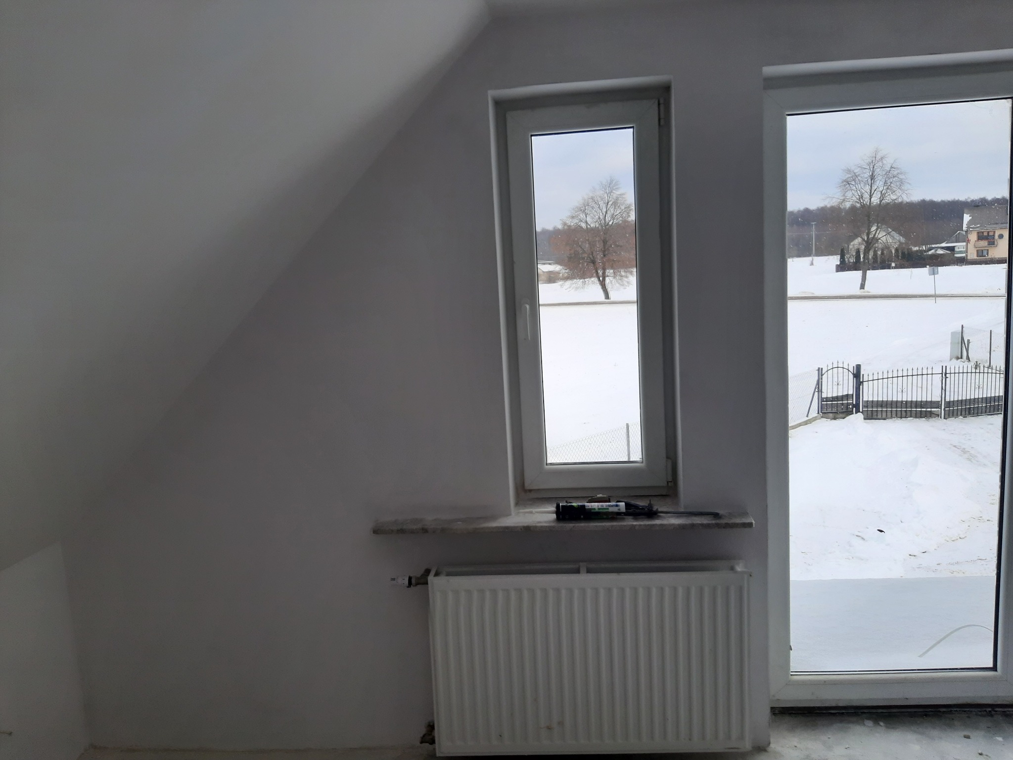 Poddasze po remoncie: białe ściany, okno z widokiem na zaśnieżony krajobraz, grzejnik pod oknem, tuba z silikonem na parapecie, duże okno balkonowe.
