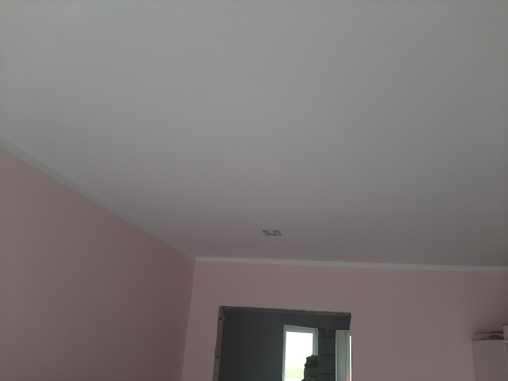 Różowe ściany z białym sufitem i listwami, widoczne niedokończone obramowanie drzwi z fragmentami muru, sufit z zamontowanym punktem oświetleniowym.