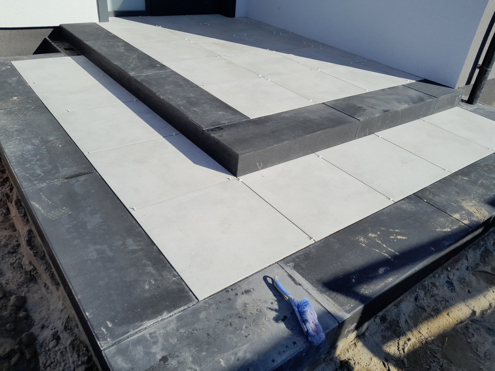 Układanie jasnych płytek na schodach wejściowych z ciemnym obramowaniem, widoczne krzyżyki dystansowe i szczotka do fugowania.