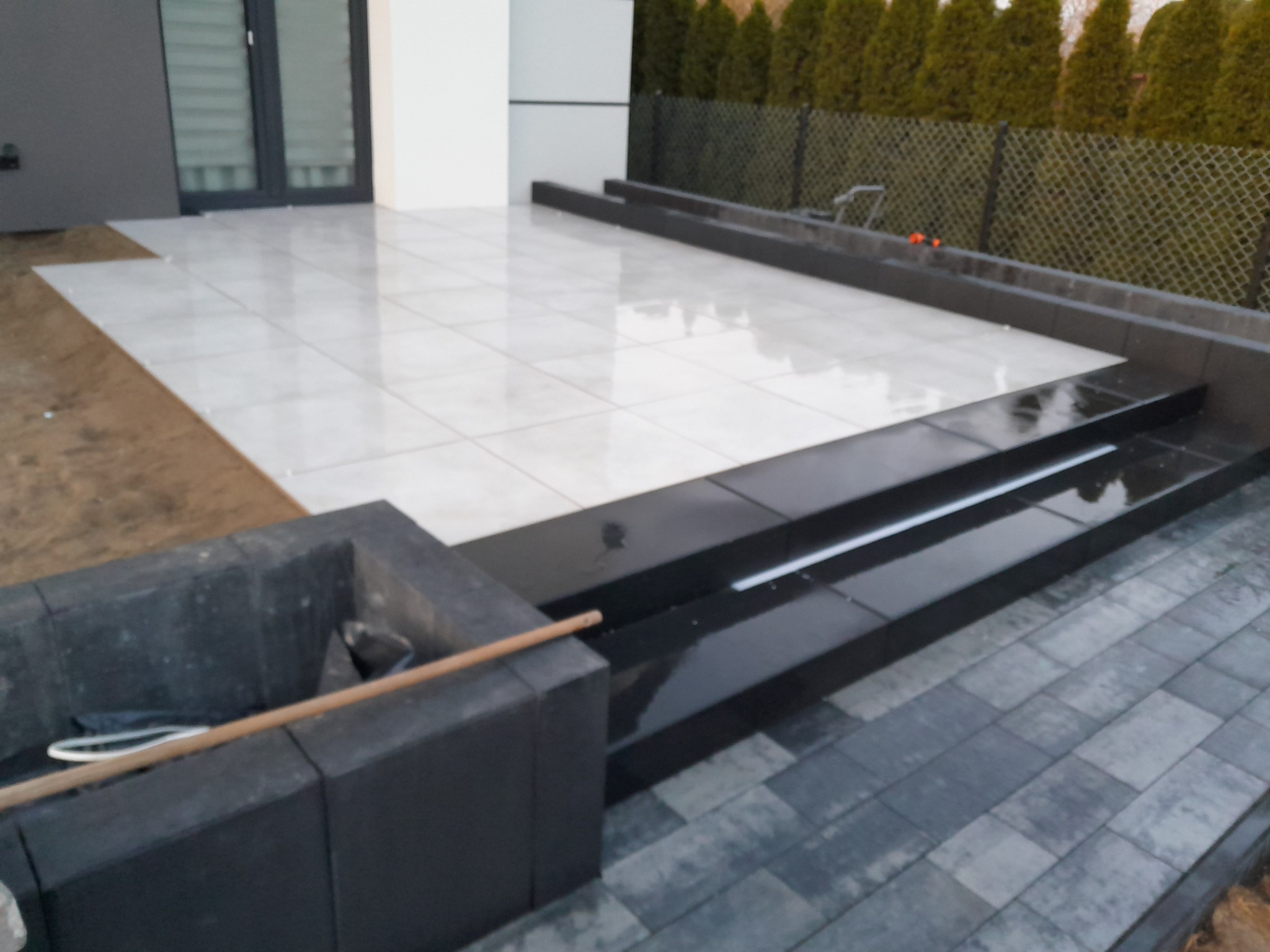 Jasne płytki tarasowe ułożone przy nowoczesnym domu, z czarnymi schodami i obrzeżami, widoczny fragment kostki brukowej i element paleniska z drewnem.