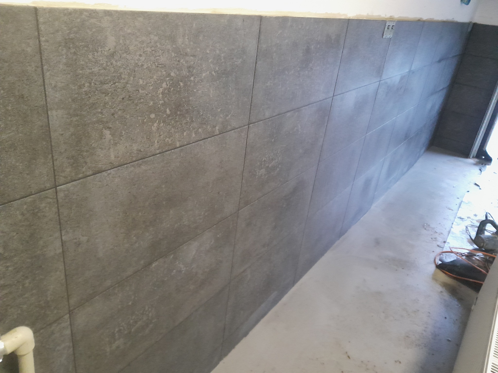 Ściana wyłożona szarymi, matowymi płytkami imitującymi kamień, z widocznymi śladami po kleju na podłodze i elementami instalacji elektrycznej na górze.