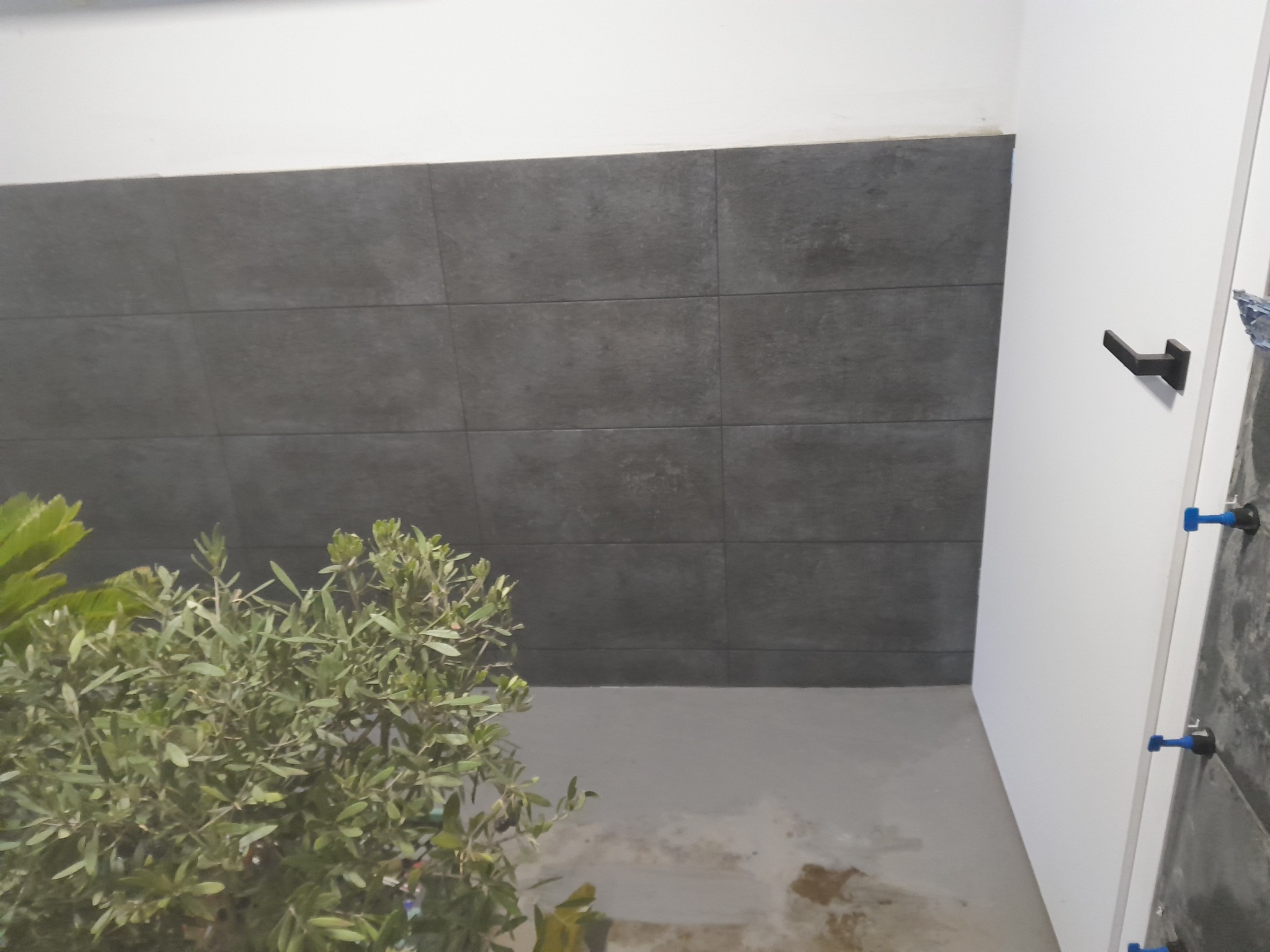Szare, matowe płytki ceramiczne ułożone na ścianie, widoczny fragment białej ściany z czarną klamką i rośliną w rogu pomieszczenia, na podłodze ślady po fugowaniu.