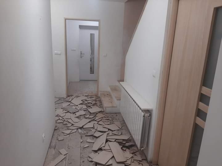 Korytarz w trakcie demontażu starej podłogi, widoczne fragmenty płytek na podłodze, przygotowanie do położenia nowej nawierzchni, drzwi wewnętrzne po prawej stronie, grzejnik z białym parapetem.