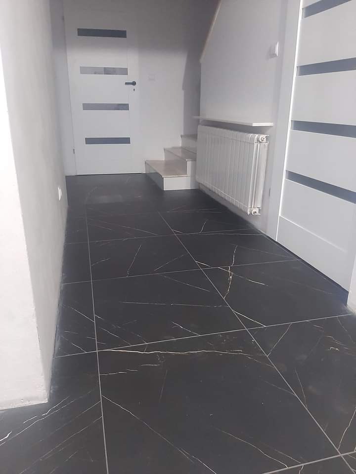 Wykończony korytarz z czarną, marmurkową podłogą, białymi drzwiami z szarymi akcentami i schodami prowadzącymi na górę, widoczny grzejnik.