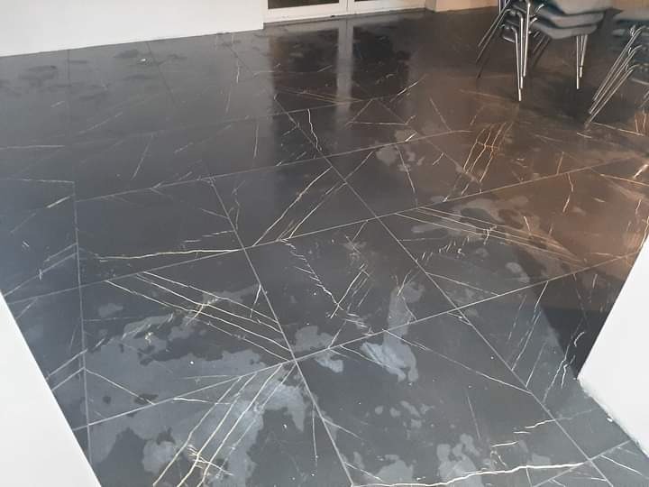 Połyskująca, czarna podłoga z płytek imitujących marmur z wyraźnymi złotymi żyłami, odbijająca światło, z widocznymi krzesłami w tle.