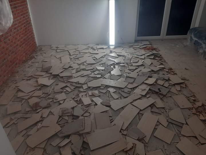 Podłoga w trakcie demontażu, pokryta fragmentami usuniętych płytek, widoczna ceglana ściana i okno w tle.
