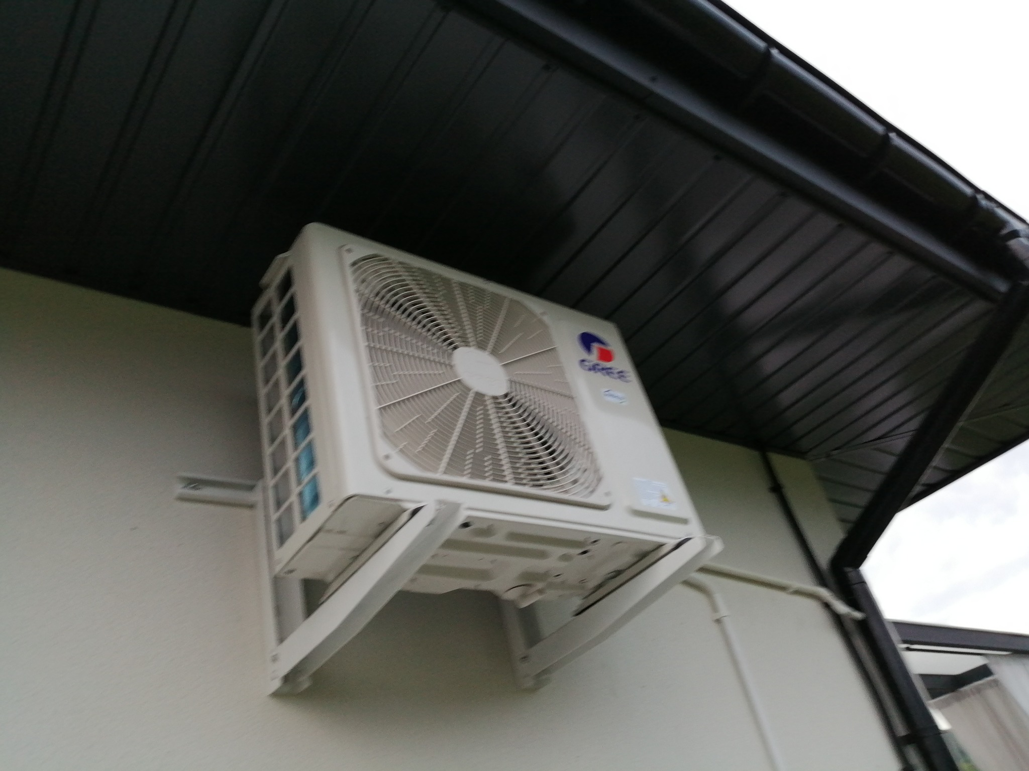 Biała jednostka zewnętrzna klimatyzatora marki Gree zamontowana na jasnej elewacji budynku, widoczna z dołu pod kątem, z fragmentem ciemnej podbitki dachowej i rynny.