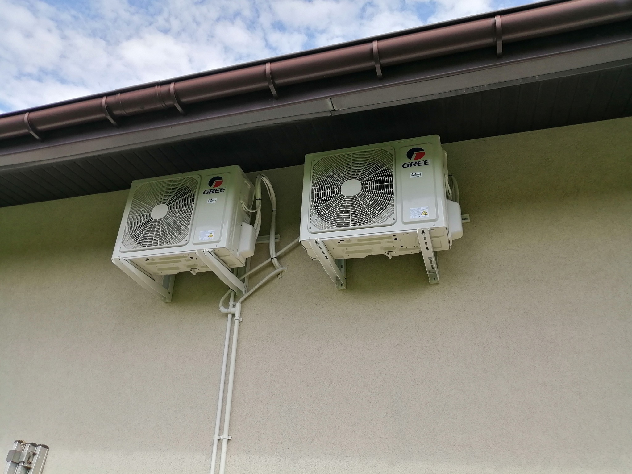 Dwa białe klimatyzatory marki Gree zamontowane na jasnej elewacji budynku, widoczne rury instalacyjne i brązowy system rynnowy.