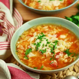 Dwa talerze zupy pomidorowej z kapustą i natką pietruszki, podane na plecionym podkładce, obok leży lniana ściereczka w biało-czerwone paski.