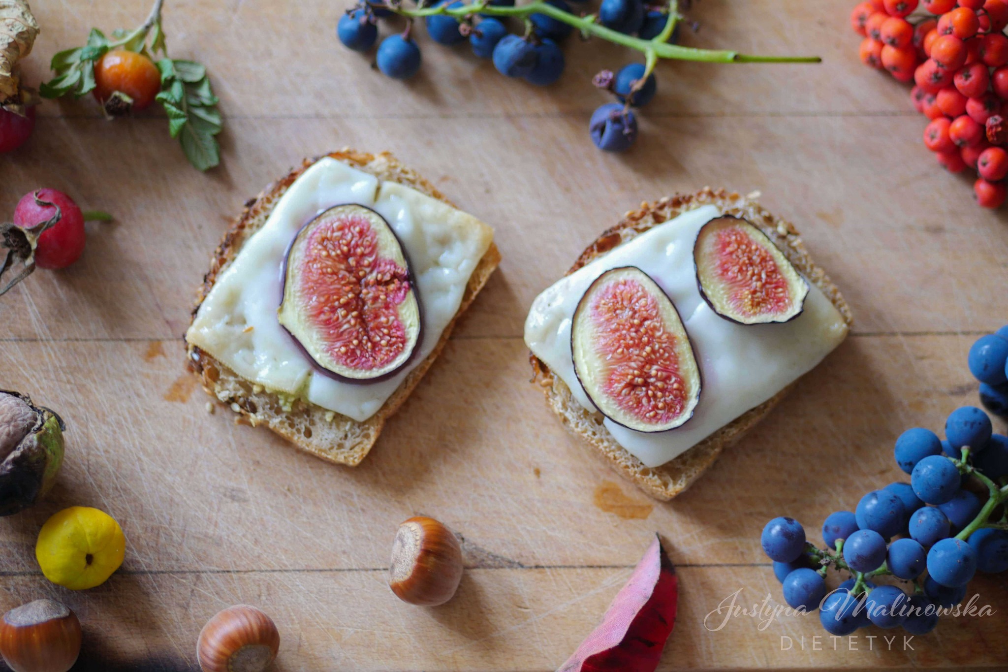 Dwa tosty z serem i figami na drewnianej desce, otoczone jesiennymi owocami: dziką różą, tarniną, jarzębiną, orzechami laskowymi, pigwą i liściem, z widocznym podpisem 'Justyna Malinowska DIETETYK'.