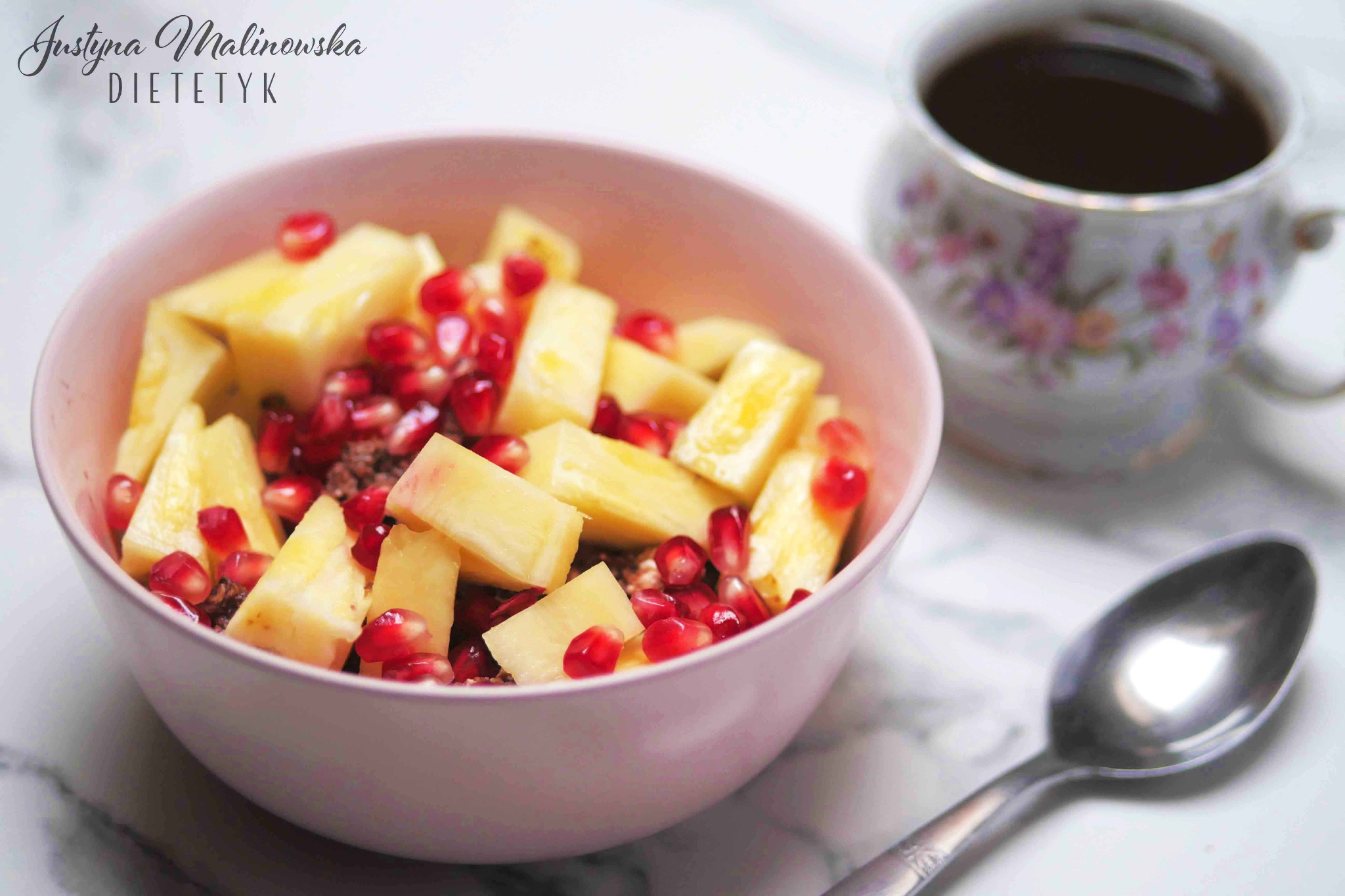 Śniadanie: kostki ananasa i granatu w różowej miseczce, obok filiżanka kawy i łyżeczka na marmurowym blacie, logo dietetyka w lewym górnym rogu.