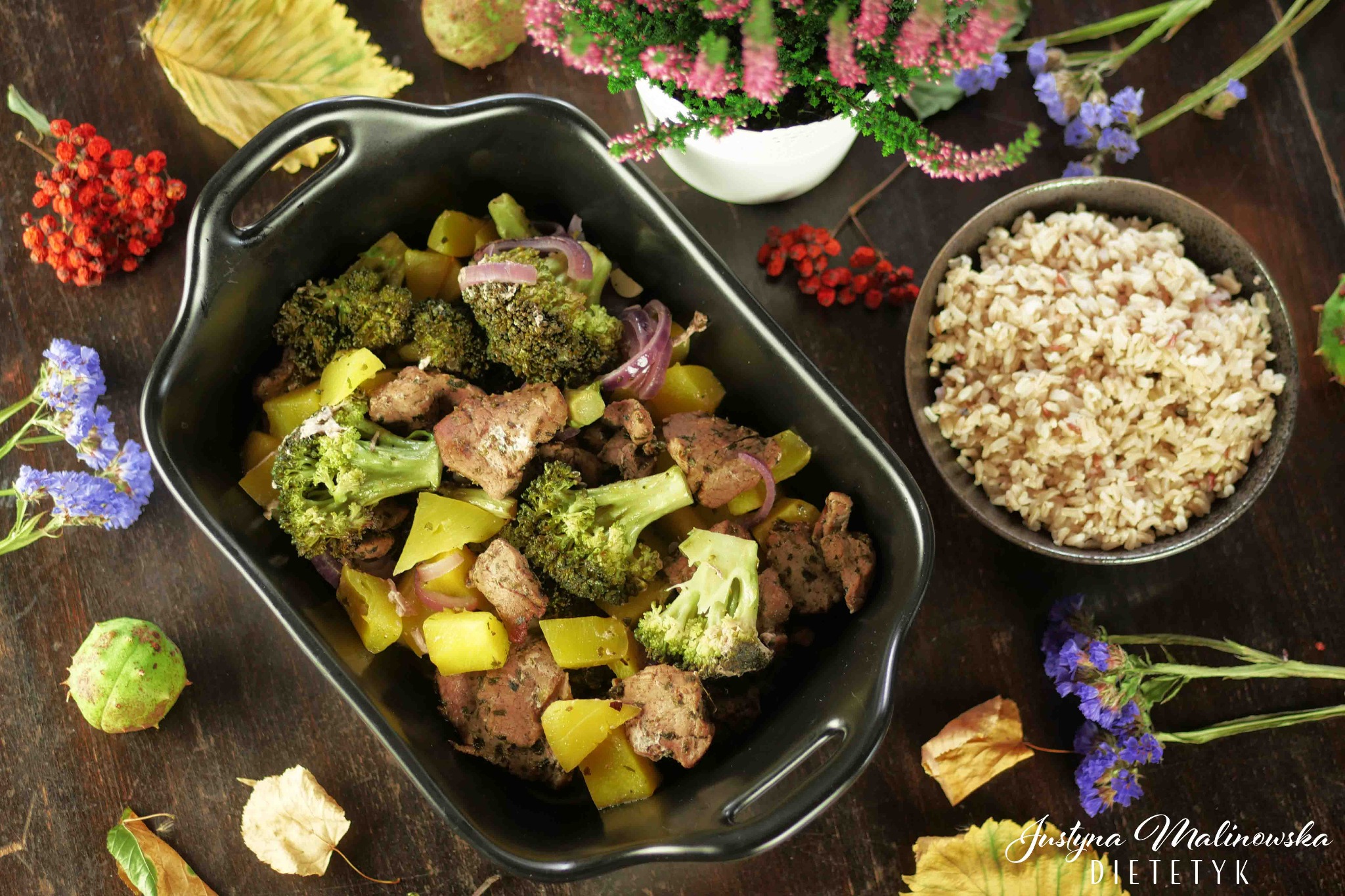 Danie z mięsem, brokułami, ziemniakami i czerwoną cebulą w czarnym naczyniu, obok miseczka brązowego ryżu, całość udekorowana kwiatami i liśćmi na drewnianym stole. W prawym dolnym rogu widoczny...