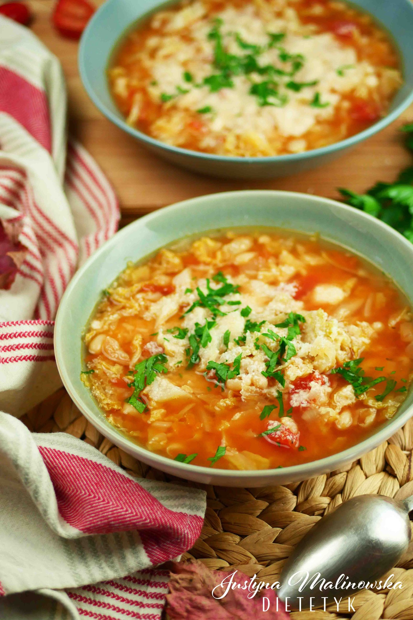 Dwa talerze zupy pomidorowej z kapustą i natką pietruszki, podane na plecionym podkładce, obok leży lniana ściereczka w biało-czerwone paski.