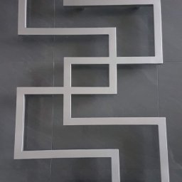 WSS - Welding Stainless Steel - Geometryczna dekoracja ścienna z profili metalowych o matowym wykończeniu, układająca się w abstrakcyjny wzór na ciemnoszarej, płytkowej ścianie.