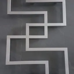 Geometryczna dekoracja ścienna z profili metalowych o matowym wykończeniu, układająca się w abstrakcyjny wzór na ciemnoszarej, płytkowej ścianie.