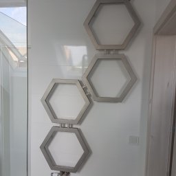 WSS - Welding Stainless Steel - Grzejnik łazienkowy dekoracyjny w kształcie trzech połączonych ze sobą heksagonów, zamontowany na białej, kafelkowanej ścianie w łazience, widoczne zawory i termostat.