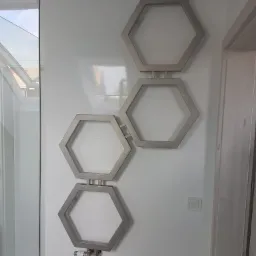 Grzejnik łazienkowy dekoracyjny w kształcie trzech połączonych ze sobą heksagonów, zamontowany na białej, kafelkowanej ścianie w łazience, widoczne zawory i termostat.