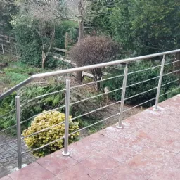 Zewnętrzna balustrada ze stali nierdzewnej na schodach prowadzących do ogrodu, widok z góry na brązowe płytki i zieloną roślinność w tle.