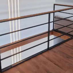 Nowoczesna balustrada metalowa z drewnianym blatem, czarne ramy, widok z góry na podłogę z paneli, w tle pionowe lampy LED.