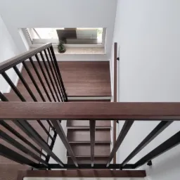 Nowoczesne schody z drewnianymi stopniami i czarną metalową balustradą, widok z góry na jasne wnętrze z oknem dachowym.